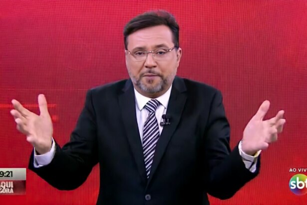 Apresentador Geraldo Luís no estúdio do programa "Aqui Agora", no SBT. Ele usa um terno preto, camisa branca, gravata listrada e óculos, com as mãos abertas e levantadas em gesticulação, em frente a um fundo de LED vermelho. O logo do SBT e a indicação "AO VIVO" aparecem no canto inferior direito da tela, enquanto no canto inferior esquerdo há um letreiro com o nome do programa