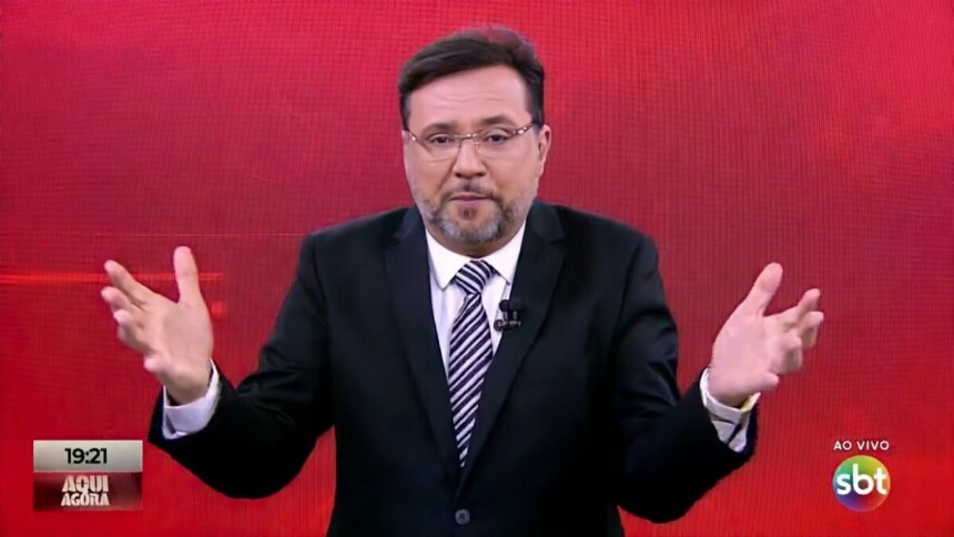 Apresentador Geraldo Luís no estúdio do programa "Aqui Agora", no SBT. Ele usa um terno preto, camisa branca, gravata listrada e óculos, com as mãos abertas e levantadas em gesticulação, em frente a um fundo de LED vermelho. O logo do SBT e a indicação "AO VIVO" aparecem no canto inferior direito da tela, enquanto no canto inferior esquerdo há um letreiro com o nome do programa