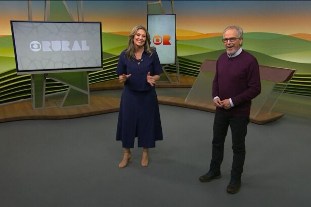 Cristina Vieira e Nélson Araújo, apresentadores do Globo Rural, em pé no estúdio do programa, que tem painéis digitais ao fundo. Cristina veste um conjunto azul marinho de blusa e saia, e Nélson usa uma camisa social clara, suéter vinho e calça preta