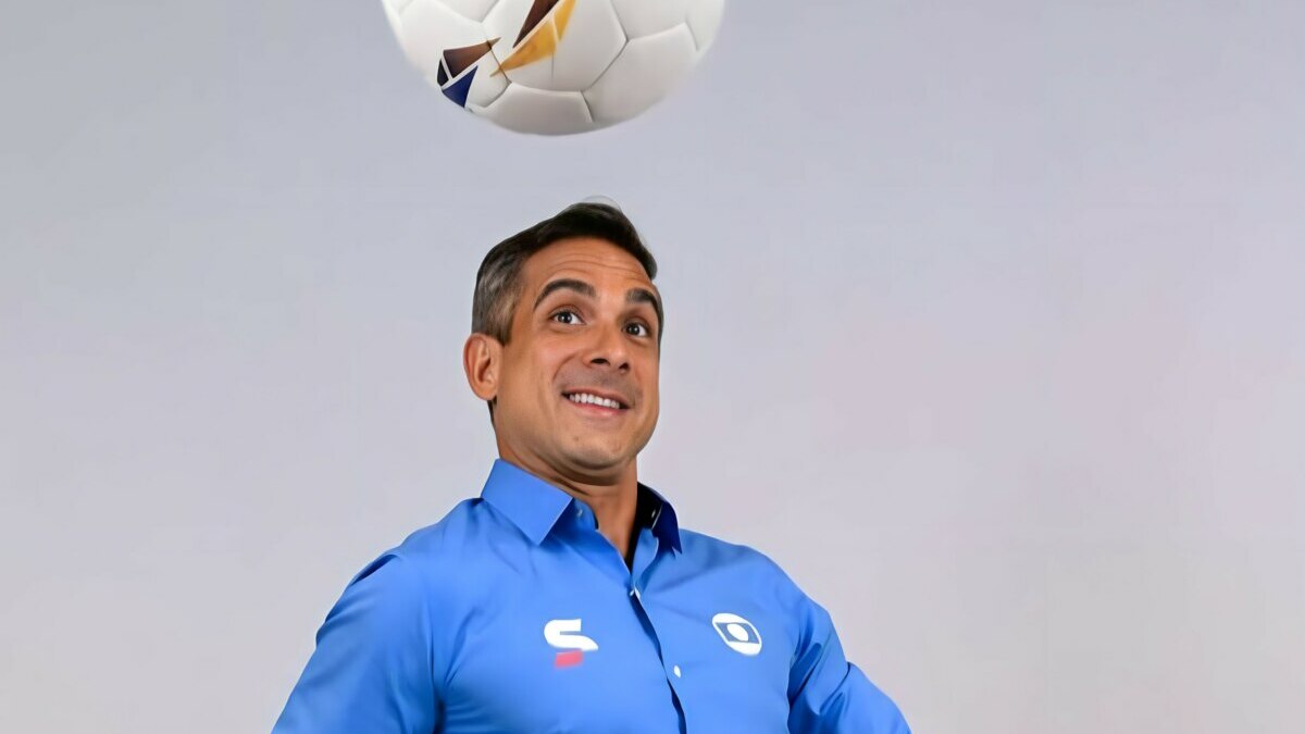 O narrador esportivo Gustavo Villani, com camisa azul, fardamento da TV Globo, olha para uma bola de futebol no ar