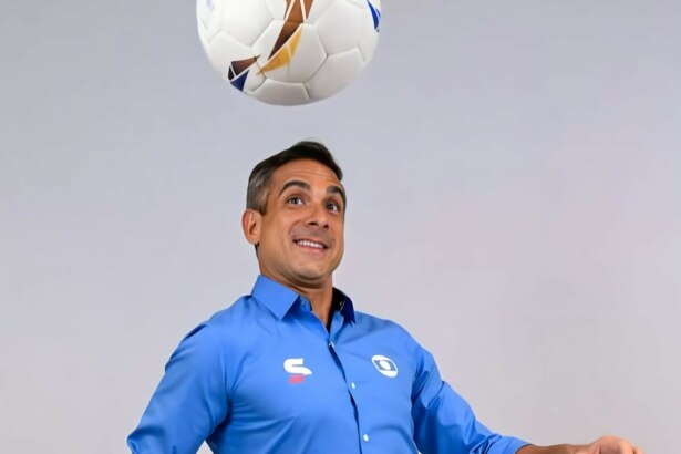 O narrador esportivo Gustavo Villani, com camisa azul, fardamento da TV Globo, olha para uma bola de futebol no ar