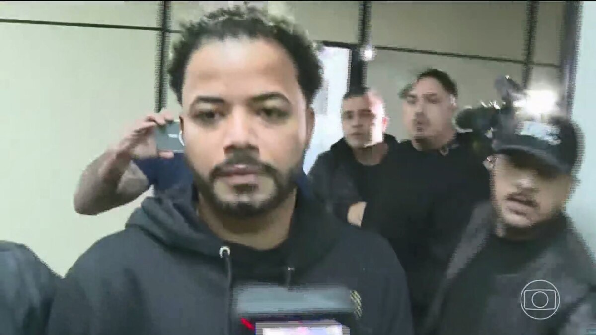 Influenciador Hytalo Santos em um ambiente interno. Ele está no centro da imagem, olhando para a câmera com uma expressão séria e usando um moletom preto. Ao seu redor, outras pessoas aparecem desfocadas, com alguns segurando celulares e câmeras com flashes. O logo da TV Globo é visível no canto inferior direito.