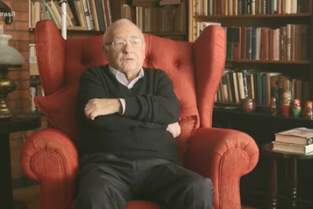 Luis Fernando Verissimo sentado em uma ampla poltrona vermelha, cercado por estante repleta de livros e itens decorativos, incluindo bonecas russas, em ambiente que remete a um escritório ou biblioteca. Cena exibida pelo *Fala Brasil* (Record) na cobertura sobre sua internação em Porto Alegre