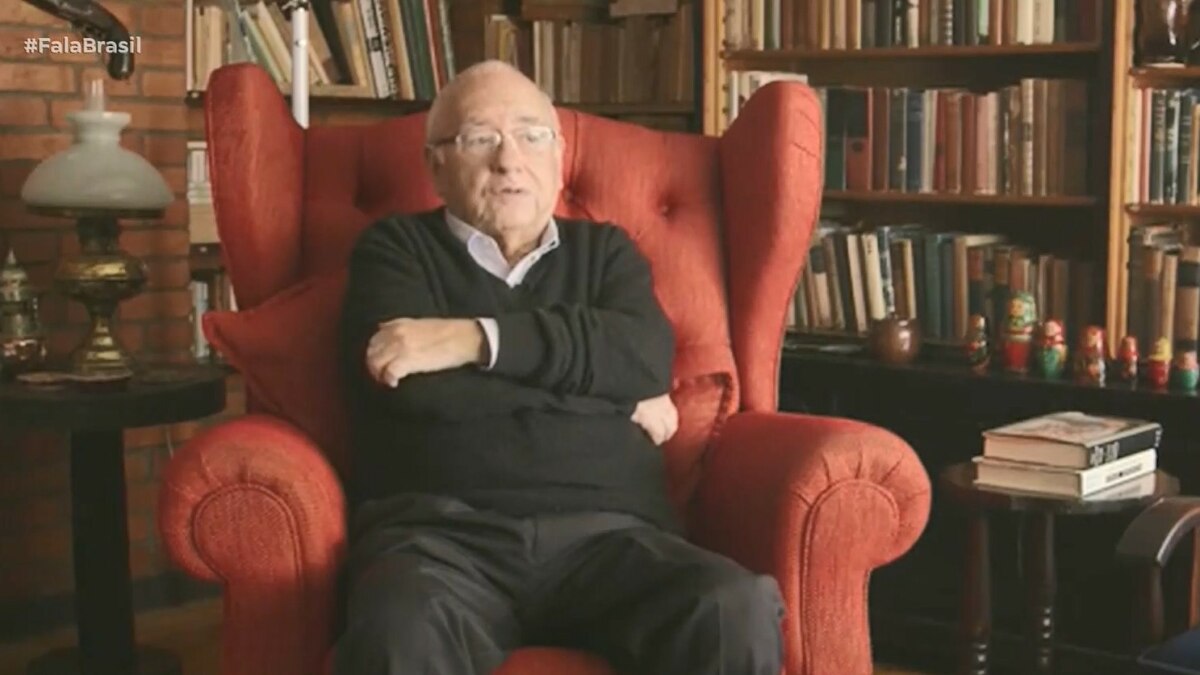 Luis Fernando Verissimo sentado em uma ampla poltrona vermelha, cercado por estante repleta de livros e itens decorativos, incluindo bonecas russas, em ambiente que remete a um escritório ou biblioteca. Cena exibida pelo *Fala Brasil* (Record) na cobertura sobre sua internação em Porto Alegre