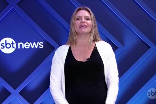 Jornalista Nathalia Fruet, do SBT News, em plantão jornalístico ao vivo. Ela está no estúdio com um fundo de LED azul escuro com o logo da emissora, usando uma blusa preta e um casaco branco, com a boca aberta, mas sem áudio
