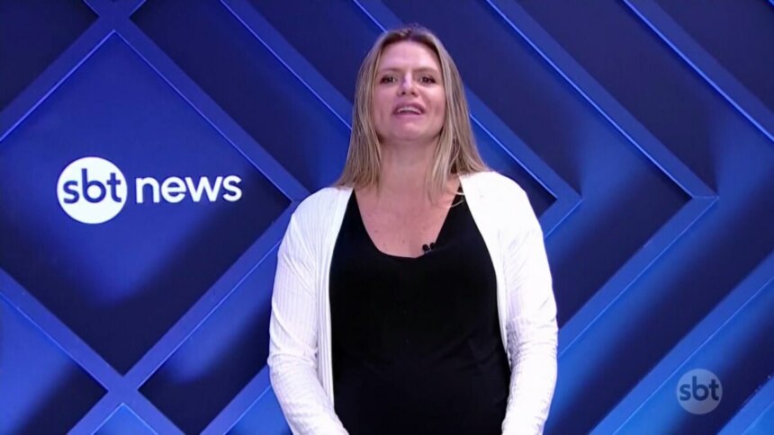 Jornalista Nathalia Fruet, do SBT News, em plantão jornalístico ao vivo. Ela está no estúdio com um fundo de LED azul escuro com o logo da emissora, usando uma blusa preta e um casaco branco, com a boca aberta, mas sem áudio