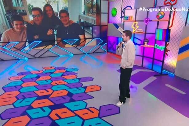 João Silva, com expressão comovida e olhar voltado para o vídeo, presta homenagem a Faustão durante a estreia do Programa do João, exibido no domingo (10) pelo SBT, em celebração ao Dia dos Pais. A tela ao fundo exibe momentos compartilhados entre pai e filho