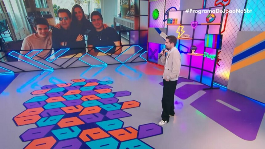 João Silva, com expressão comovida e olhar voltado para o vídeo, presta homenagem a Faustão durante a estreia do Programa do João, exibido no domingo (10) pelo SBT, em celebração ao Dia dos Pais. A tela ao fundo exibe momentos compartilhados entre pai e filho