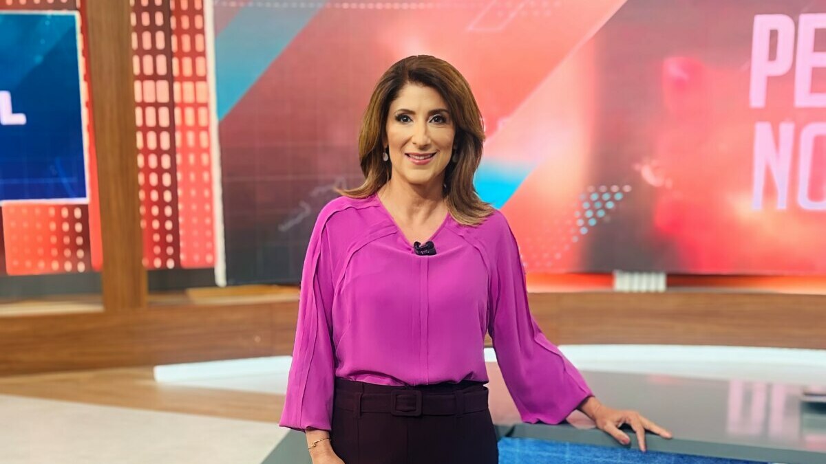 A jornalista Simone Queiroz em pé em um estúdio de telejornal do sBT. Ela usa uma blusa magenta e calças escuras, sorri para a câmera com a mão direita apoiada em uma bancada com o logo do SBT. Ao fundo, um painel digital exibe gráficos e o texto "PE NO".