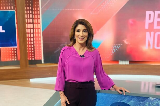 A jornalista Simone Queiroz em pé em um estúdio de telejornal do sBT. Ela usa uma blusa magenta e calças escuras, sorri para a câmera com a mão direita apoiada em uma bancada com o logo do SBT. Ao fundo, um painel digital exibe gráficos e o texto "PE NO".