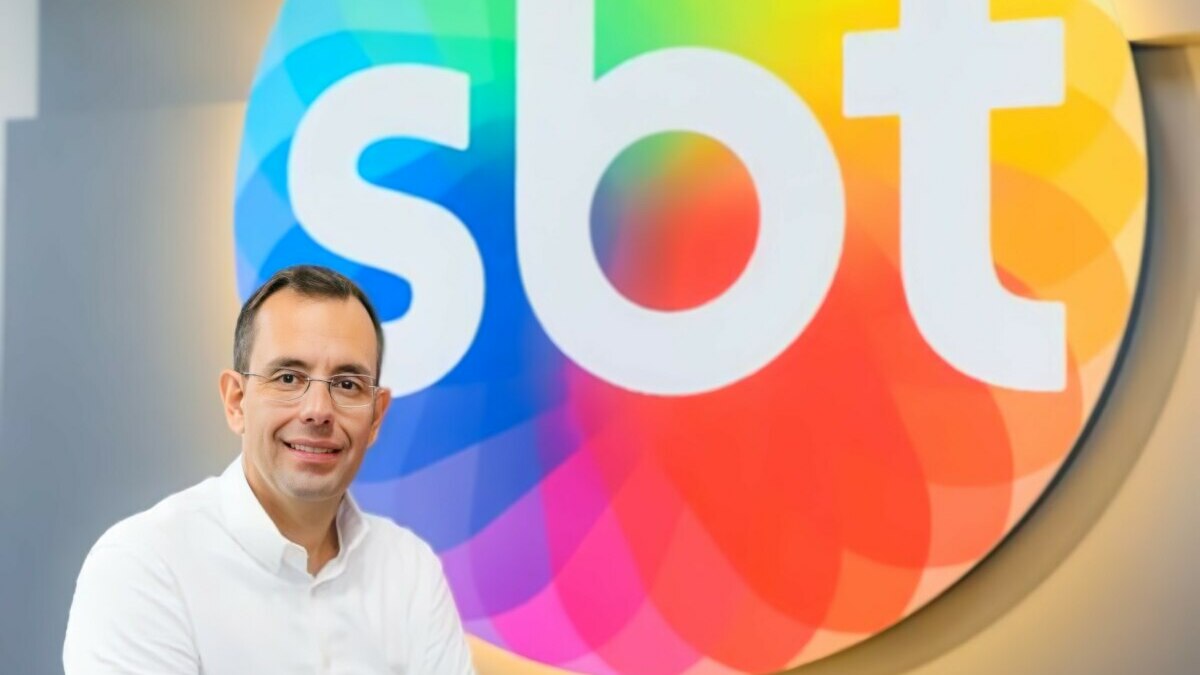 O executivo Vicente Varela posa sorrindo, com os braços cruzados, em frente ao logo colorido do SBT.