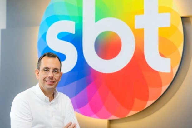 O executivo Vicente Varela posa sorrindo, com os braços cruzados, em frente ao logo colorido do SBT.