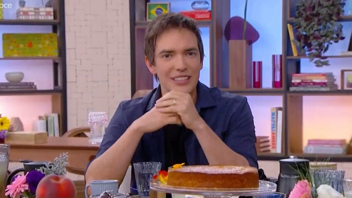 O jornalista Alessandro Jodar sorri na mesa do Mais Você, que está repleta de comida e um bolo. Ele usa uma camisa azul com uma camiseta preta.