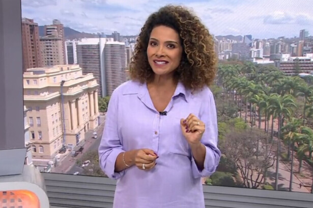 A jornalista Aline Aguiar, de blusa lilás, sorri para a câmera e aponta para sua barriga de grávida no cenário do telejornal MG1, com a cidade de Belo Horizonte ao fundo.