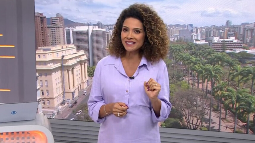 A jornalista Aline Aguiar, de blusa lilás, sorri para a câmera e aponta para sua barriga de grávida no cenário do telejornal MG1, com a cidade de Belo Horizonte ao fundo.