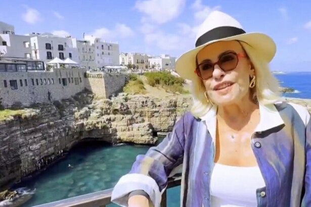 Ana Maria Braga de chapéu branco e óculos de sol, sorrindo e usando uma blusa colorida com listras, em uma vista deslumbrante de um vilarejo à beira-mar na Itália, em vídeo exibido no Mais Você