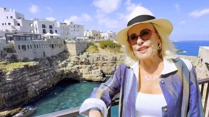 Ana Maria Braga de chapéu branco e óculos de sol, sorrindo e usando uma blusa colorida com listras, em uma vista deslumbrante de um vilarejo à beira-mar na Itália, em vídeo exibido no Mais Você