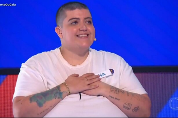 Ana Vilela, de cabeça raspada, sorrindo e olhando para cima com as mãos entrelaçadas sobre o peito, no cenário azul e vermelho do programa Acerte ou Caia.