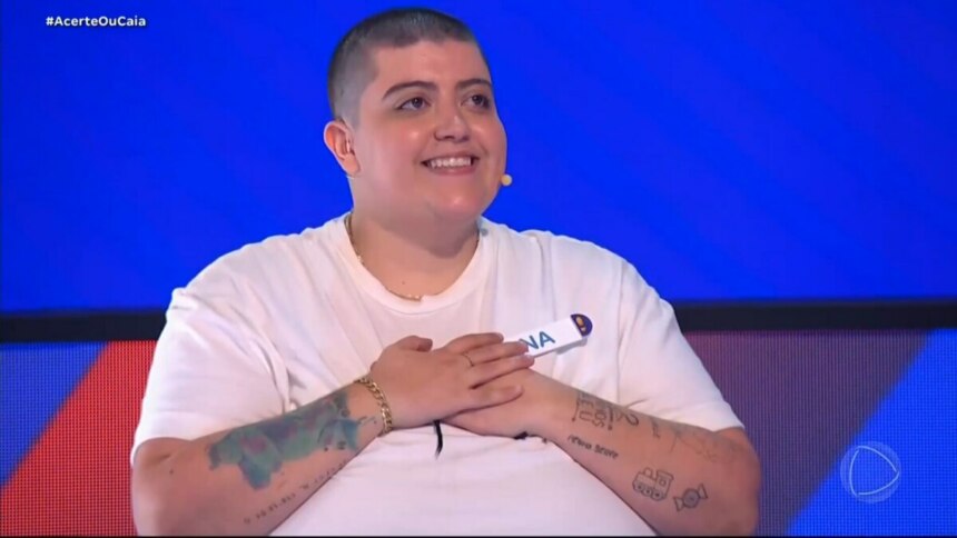 Ana Vilela, de cabeça raspada, sorrindo e olhando para cima com as mãos entrelaçadas sobre o peito, no cenário azul e vermelho do programa Acerte ou Caia.