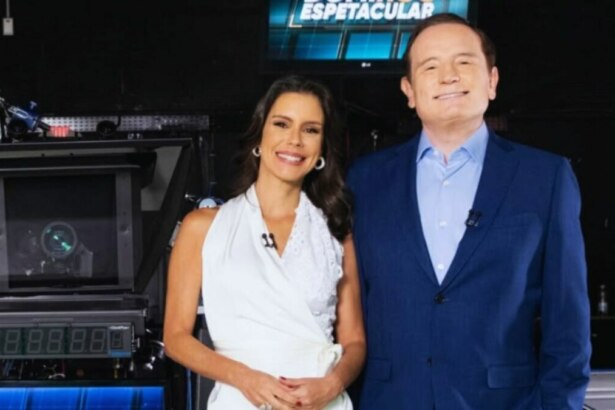 Camila Busnello e Roberto Cabrini posam sorrindo nos bastidores do Domingo Espetacular. Ela veste um vestido branco e ele um terno azul.