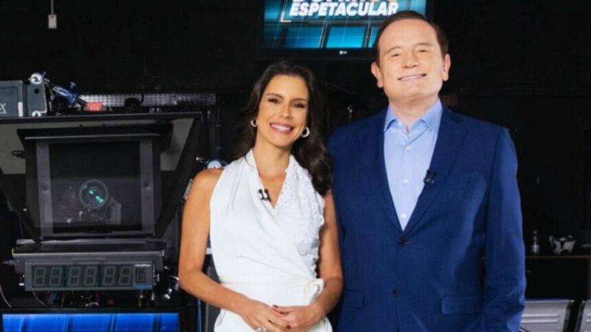 Camila Busnello e Roberto Cabrini posam sorrindo nos bastidores do Domingo Espetacular. Ela veste um vestido branco e ele um terno azul.