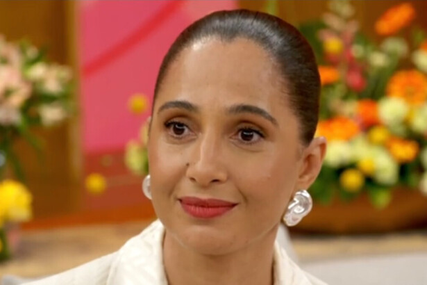 A atriz Camila Pitanga, com o cabelo preso em um coque, usa grandes brincos brancos. Ela está sorrindo discretamente em uma participação no programa Encontro, com um fundo de flores borrado.