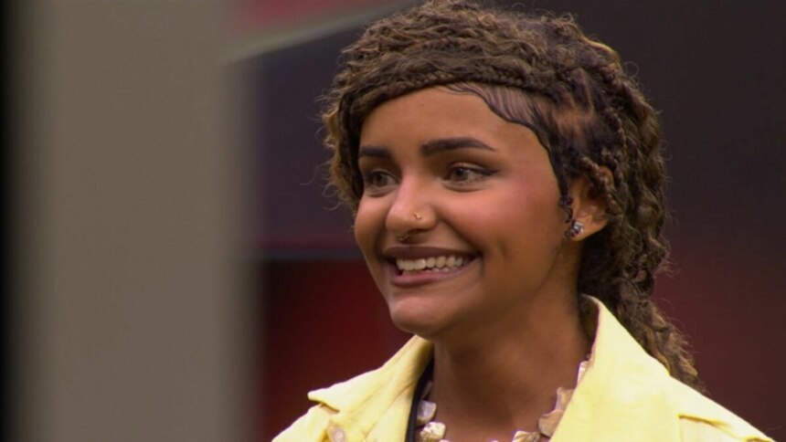 Camille Vitória, de cabelos trançados, sorrindo e olhando para o lado, com uma jaqueta amarela em trecho da Prova do Jingle do programa Estrela da Casa.