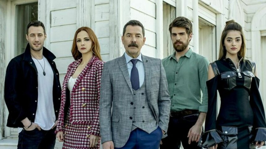 Os atores da novela Cruel Istambul (Fikret Kuskan, Deniz Ugur, Berker Guven, Mine Tugay e Sera Kutlubey) estão posando para a foto, sérios e alinhados, em frente a uma casa branca.