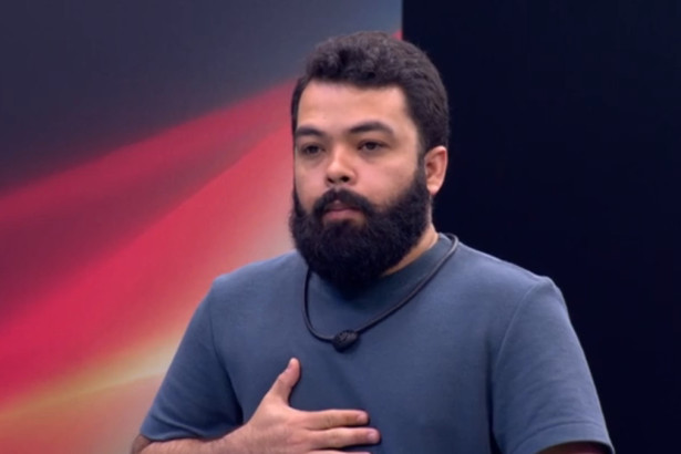 O participante Daniel Sobral, barbudo, com uma camisa azul, gesticulando no peito, no cenário da Prova do Jingle do reality show Estrela da Casa.