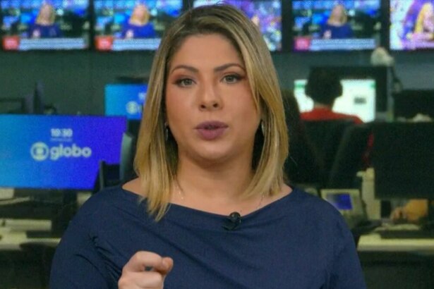 Daniela Lima com cabelos loiros e curtos, usando uma blusa azul-marinho, olhando para a câmera e gesticulando com a mão direita no estúdio da GloboNews, com monitores ao fundo.