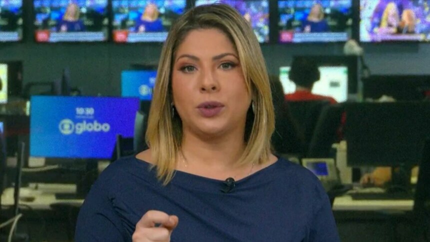Daniela Lima com cabelos loiros e curtos, usando uma blusa azul-marinho, olhando para a câmera e gesticulando com a mão direita no estúdio da GloboNews, com monitores ao fundo.