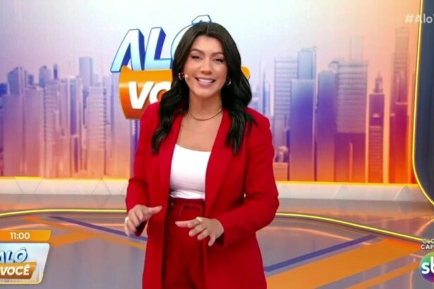 Daniele Brandi, de blazer e calça vermelhos, em pé no cenário do programa Alô Você, sorrindo e olhando para a câmera enquanto gesticula com as mãos. O logo da atração e do SBT estão na tela.