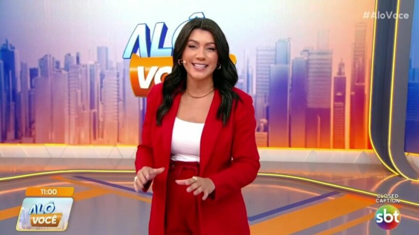 Daniele Brandi, de blazer e calça vermelhos, em pé no cenário do programa Alô Você, sorrindo e olhando para a câmera enquanto gesticula com as mãos. O logo da atração e do SBT estão na tela.