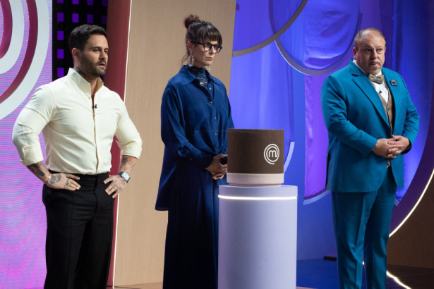 Os jurados do MasterChef Confeitaria, Diego Lozano, Helena Rizzo e Erick Jacquin, posam sérios ao lado de um objeto cilíndrico marrom, no estúdio do programa.