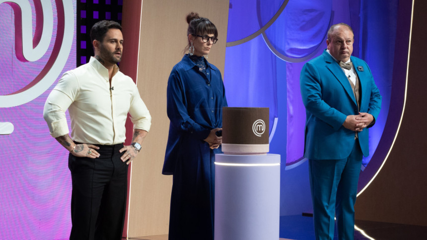 Os jurados do MasterChef Confeitaria, Diego Lozano, Helena Rizzo e Erick Jacquin, posam sérios ao lado de um objeto cilíndrico marrom, no estúdio do programa.