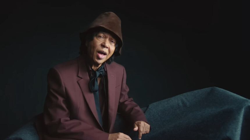 Djavan de terno vinho, gravata preta, sentado em um cenário escuro e cantando Um Brinde