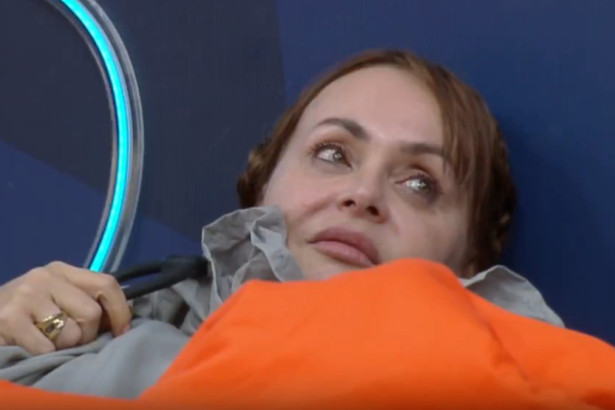 A atriz Gabriela Spanic deitada, com a cabeça apoiada em um travesseiro e coberta por uma manta laranja em A Fazenda 17. Ela parece pensativa ou triste.