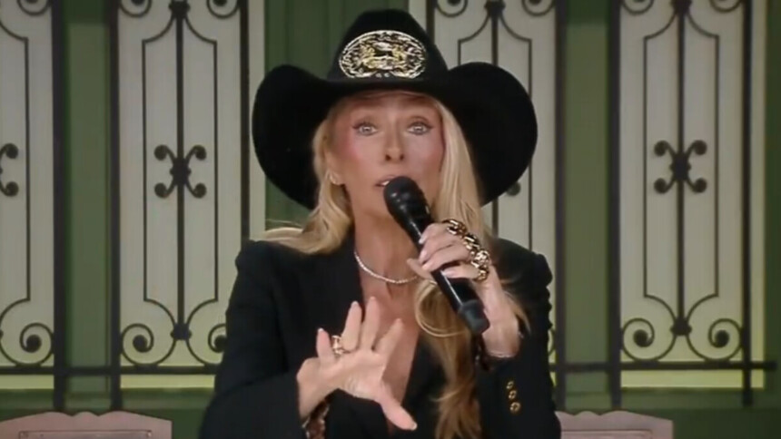 A apresentadora Adriane Galisteu, de terno preto e chapéu de caubói, segura um microfone e gesticula com a mão, em uma cena do reality show A Fazenda.
