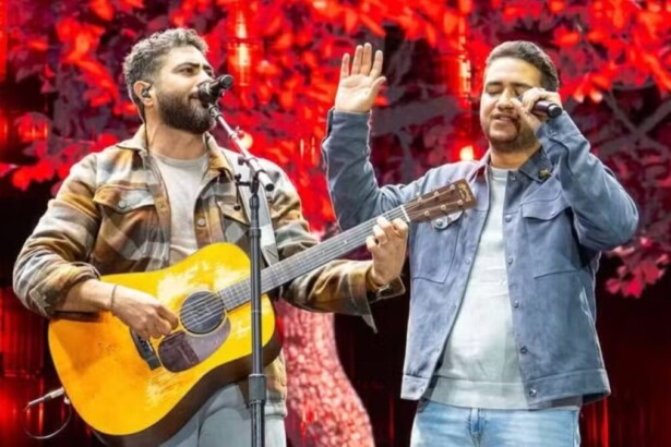 A dupla sertaneja Henrique e Juliano se apresenta em um show. Henrique, à esquerda, usa uma jaqueta xadrez e toca violão enquanto canta. Juliano, à direita, veste uma jaqueta jeans azul-clara e canta em um microfone com a mão levantada.