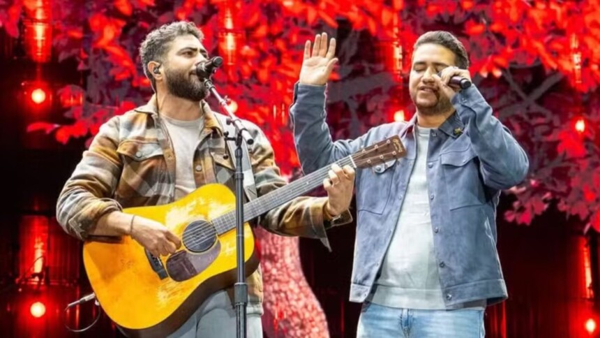A dupla sertaneja Henrique e Juliano se apresenta em um show. Henrique, à esquerda, usa uma jaqueta xadrez e toca violão enquanto canta. Juliano, à direita, veste uma jaqueta jeans azul-clara e canta em um microfone com a mão levantada.
