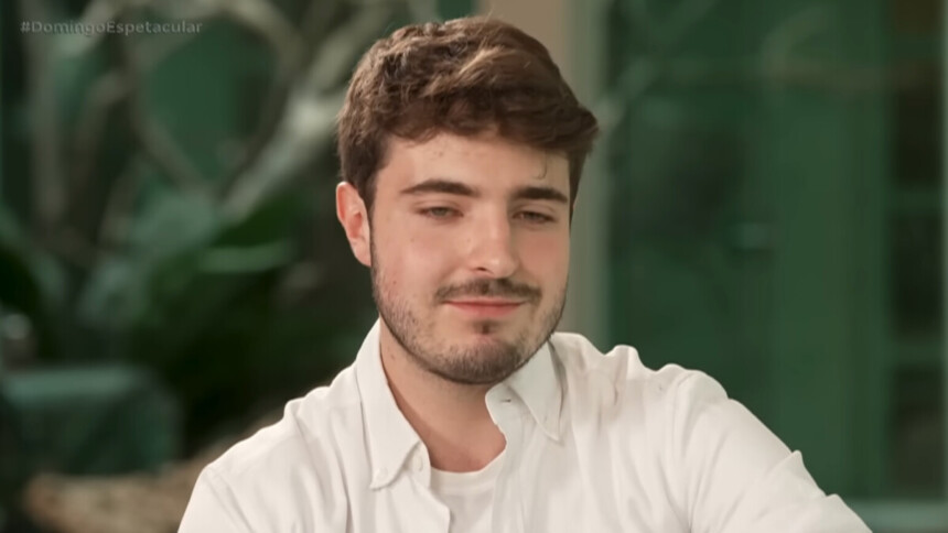 João Augusto Liberato, de barba, bigode e cabelos castanhos claros, usa uma camisa branca social e sorri de leve em uma entrevista para o programa Domingo Espetacular, com a hashtag do programa no canto superior da tela.