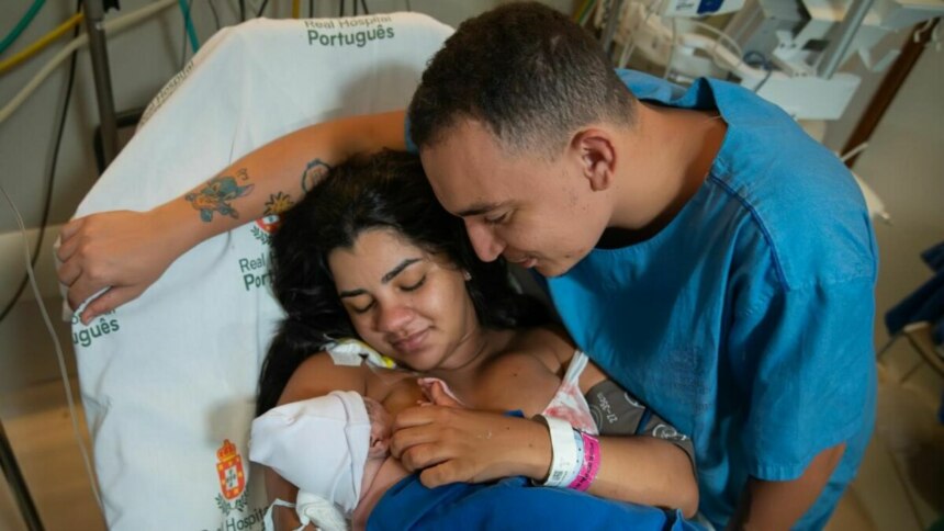 João Gomes e Ary Mirelle, emocionados, segurando o bebê recém-nascido no colo de Ary em um quarto de hospital.
