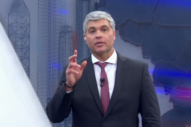 Joel Datena, com cabelos e barba grisalhos, usa um terno escuro, camisa branca e gravata vermelha. Ele está no estúdio do programa Brasil Urgente, olhando diretamente para a câmera e gesticulando com a mão direita.