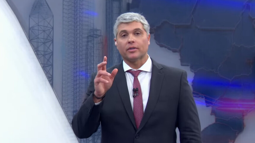 Joel Datena, com cabelos e barba grisalhos, usa um terno escuro, camisa branca e gravata vermelha. Ele está no estúdio do programa Brasil Urgente, olhando diretamente para a câmera e gesticulando com a mão direita.