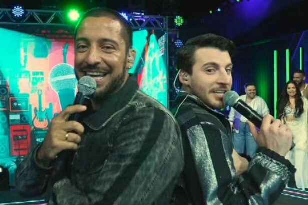 Juceir Jr e Gabriel Smaniotto, participantes do Estrela da Casa, posam sorrindo de costas um para o outro, segurando microfones no palco.