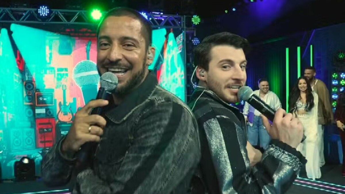 Juceir Jr e Gabriel Smaniotto, participantes do Estrela da Casa, posam sorrindo de costas um para o outro, segurando microfones no palco.
