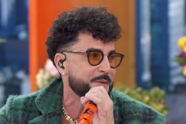 O cantor Latino, usando uma jaqueta verde peluda e óculos escuros com armação amarela, está segurando um microfone laranja com seu nome e sorrindo em uma entrevista no Encontro.