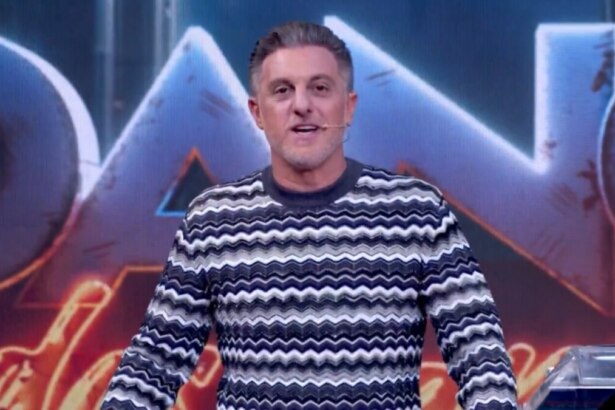 Luciano Huck de pé no palco do Domingão, com um suéter de listras brancas, pretas e cinzas, olhando para a câmera e sorrindo.