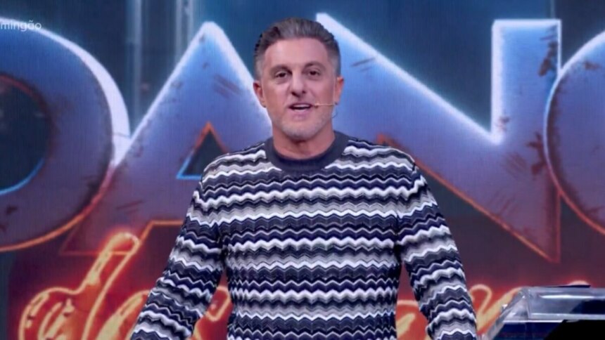 Luciano Huck de pé no palco do Domingão, com um suéter de listras brancas, pretas e cinzas, olhando para a câmera e sorrindo.