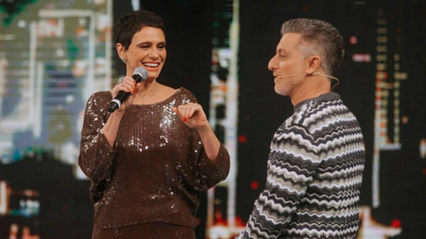 Malu Galli sorrindo e segurando microfone ao lado de Luciano Huck, ambos no palco do Domingão.